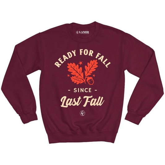 A maroon crewneck sweatshirt with the text 'READY FOR FALL SINCE LAST FALL' printed on the front, displayed against a backdrop of a cloudy sky and autumn leaves.