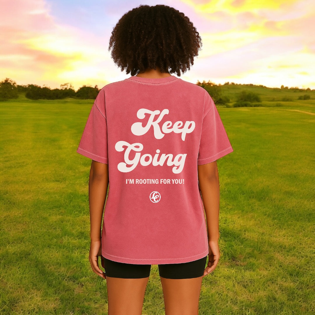 Person wearing a pink 'Keep Going' t-shirt in a grassy field with a sunset.