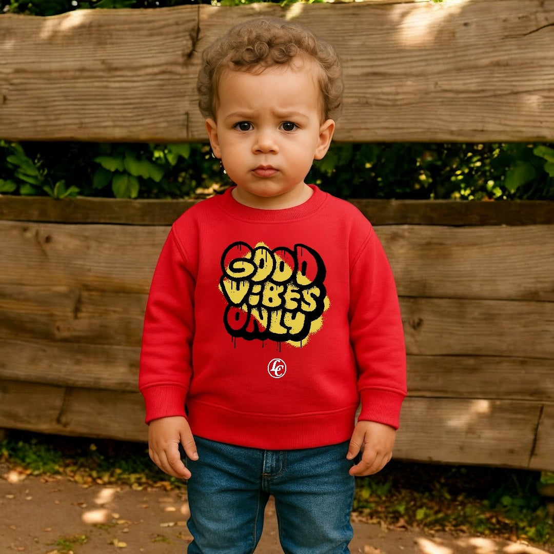 Child wearing a red sweatshirt with 'Good Vibes Only' text standing in front of a wooden fence.