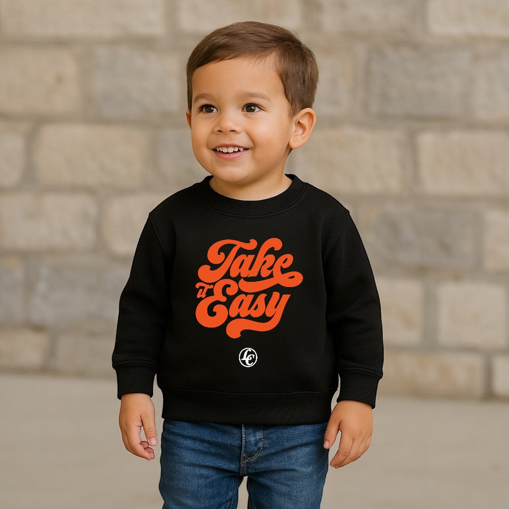 Child wearing a black sweatshirt with orange text standing against a stone wall.