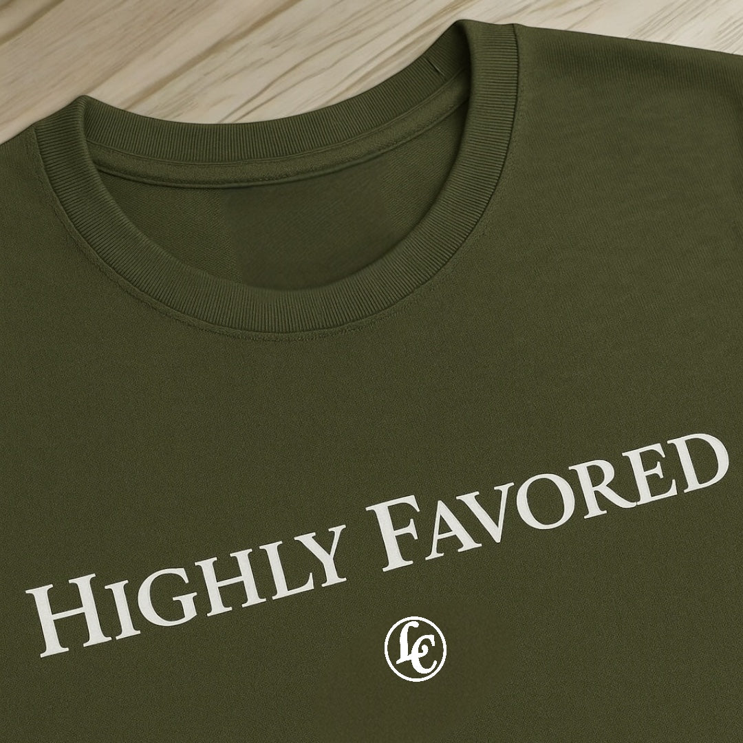 Green t-shirt with 'HIGHLY FAVORED' text and logo on a wooden surface